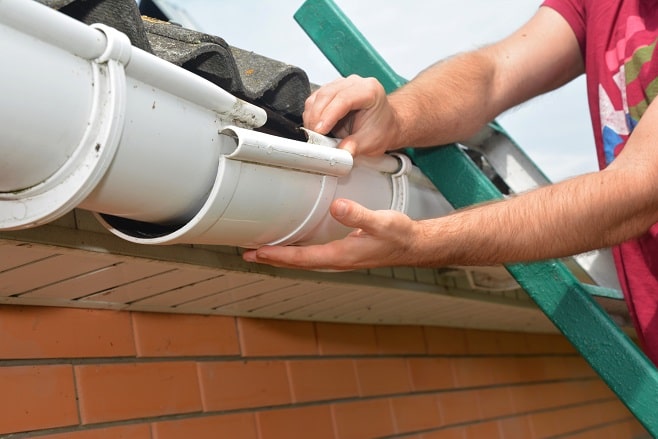 Réparation d'une gouttière qui fuit en PVC par un particulier sur une échelle, en train de reboîter un élément désolidarisé sous le bord de toit d'une maison en briques.